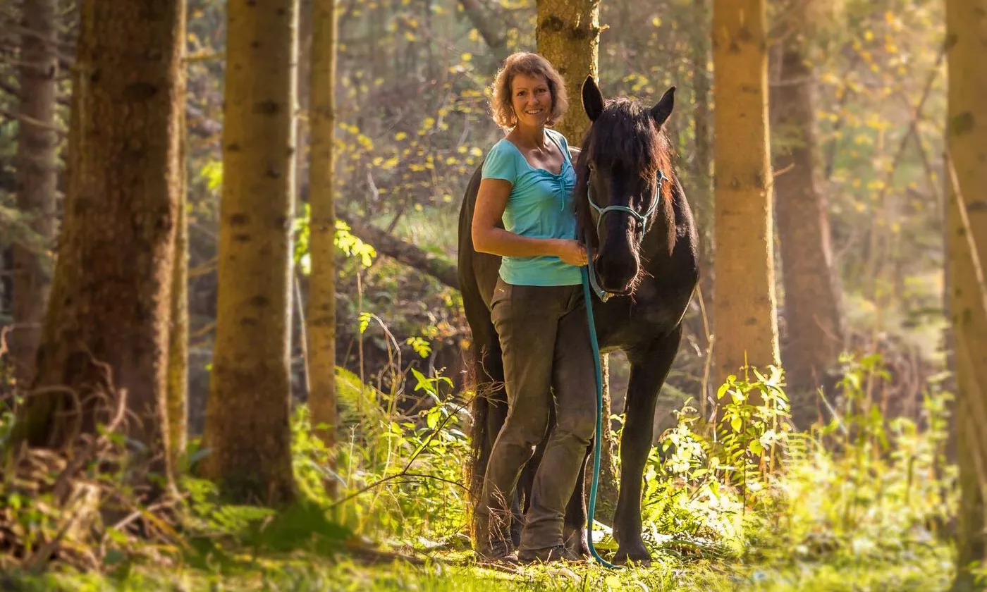 Frau mit schwarzem Pferd im sonnendurchfluteten Wald